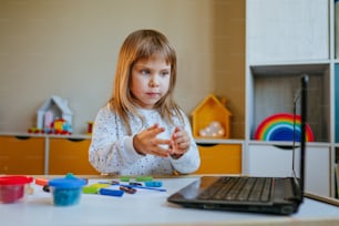 カラフルな粘土を成形する小さな女の子は、屋内のラップトップでオンライン学習レッスンを見ています。遠隔ホーム学習のコンセプト。