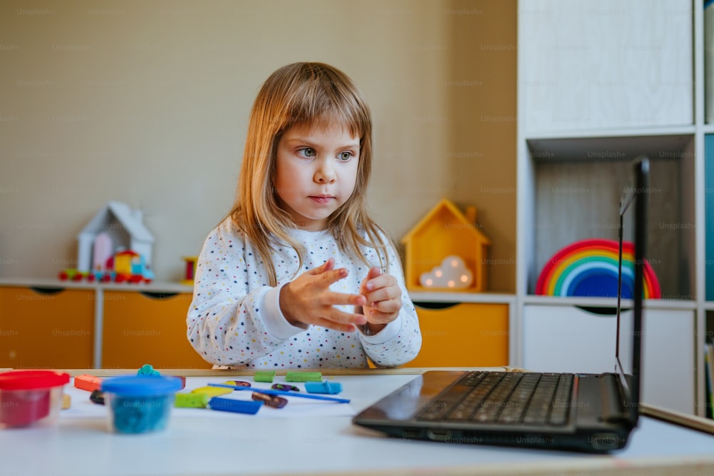 Kleines Mädchen formt bunten Ton und schaut sich Online-Lernstunde auf dem Laptop drinnen an. Fernunterrichtskonzept.