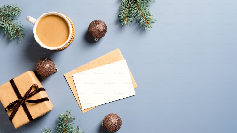 クリスマスの構図。青い背景に平らな空白のグリーティングカードのモックアップ、ギフトボックス、コーヒーカップ、茶色のボール、松の木の枝を置きます。