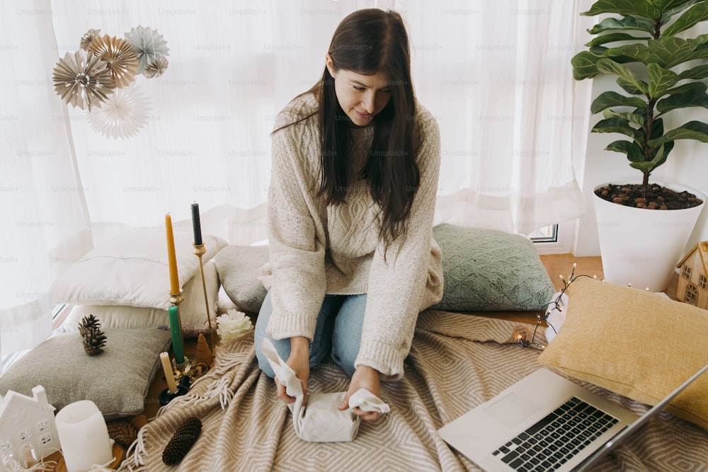 Stylish woman in cozy sweater wrapping gift box in linen fabric in festive decorated boho room and looking at laptop. Young female looking at furoshiki gift wrapping tutorial online