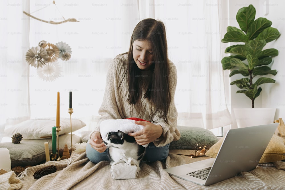 Stylish woman in cozy sweater putting santa hat on cute cat at christmas furoshiki gift and laptop in festive decorated boho room. Young female playing with furry friend. Happy Holidays!
