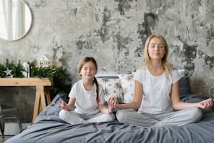 Mãe calma medita com sua filha sorridente, sentada na cama, passando o dia juntos em um quarto aconchegante. Família de pijama em casa. Conceito de práticas zen conjuntas