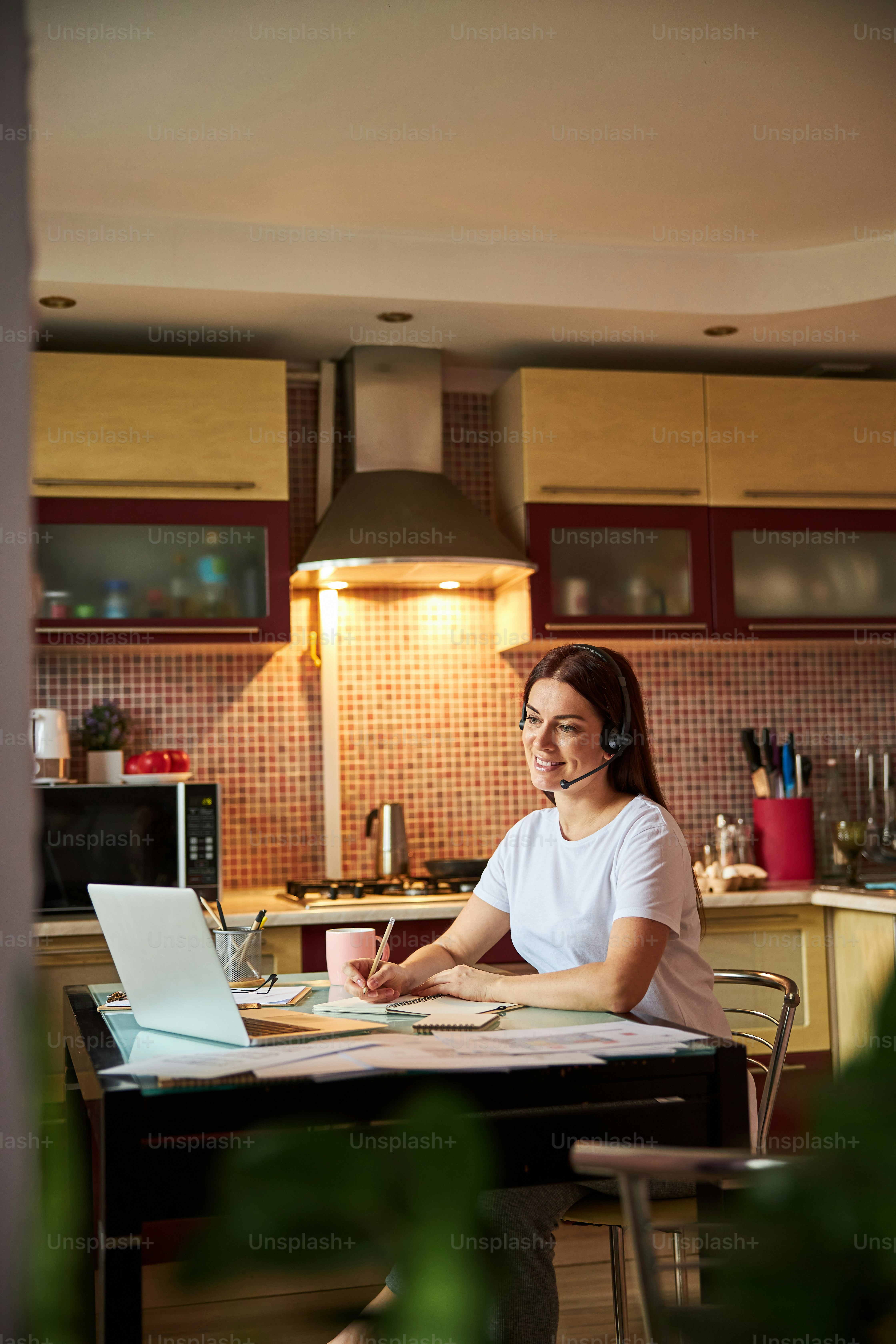 Adult woman using headphones and taking part at an online conference while writing down the necessary information