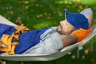 Giovane costruttore maschio pacifico che indossa l'uniforme che fa una pausa, sdraiato su un'amaca all'aperto con gli occhi chiusi in una giornata di sole. Costruzione, professione, concetto di riposo