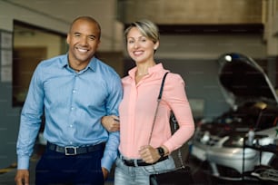 Happy African American man and his Caucasian wife standing in a car service workshop and looking at camera.