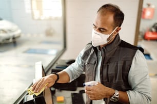 Mecánico de automóviles limpiando la computadora de la oficina con desinfectante y usando mascarilla debido a la pandemia de COVID-19.