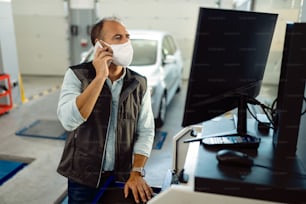 Vorarbeiter der Autowerkstatt, der über ein Mobiltelefon kommuniziert und an einem Computer arbeitet, während er aufgrund der Coronavirus-Pandemie eine Schutzmaske trägt.