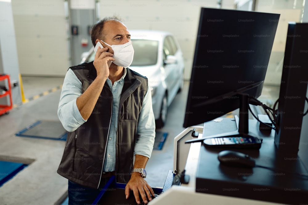 Foreman of auto repair shop communicating on mobile phone and working on a computer while wearing protective face mask due to coronavirus pandemic.