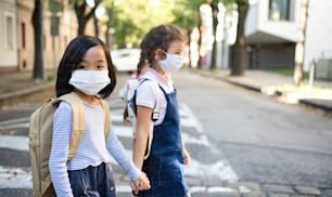 Small school girls with face mask walking outdoors in town, coronavirus concept.