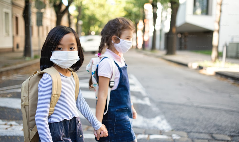 Small school girls with face mask walking outdoors in town, coronavirus concept.