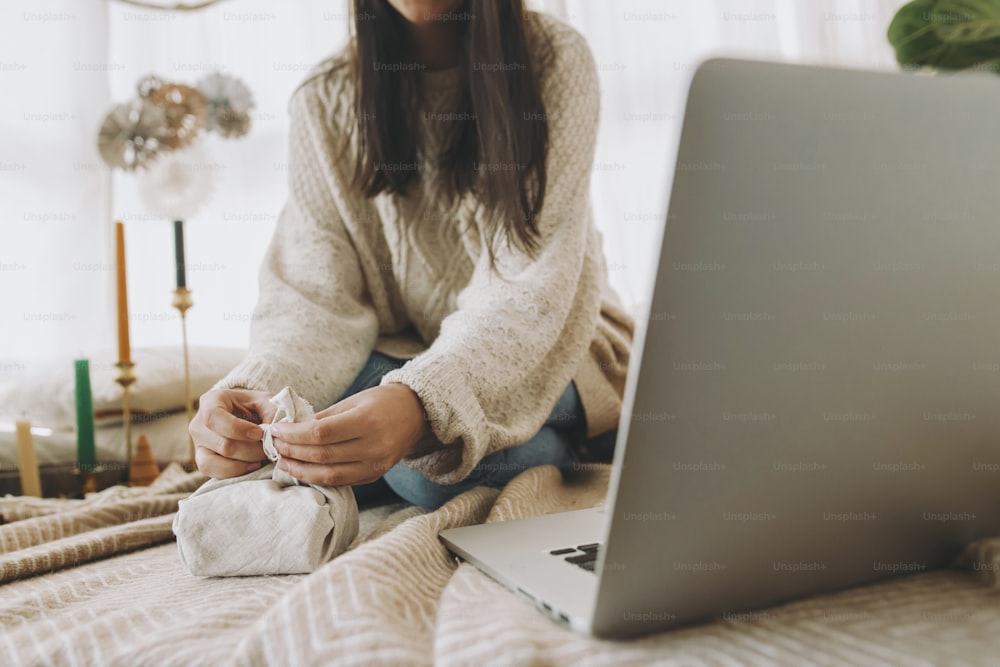 Furoshiki gift wrapping. Female wrapping christmas gift in linen fabric close up in festive decorated boho room with laptop, hands close up. Zero waste eco friendly holidays