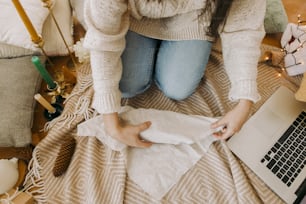 Stylish woman in cozy sweater wrapping gift box in linen fabric in festive decorated boho room with laptop, top view. Young female looking at furoshiki gift wrapping tutorial online