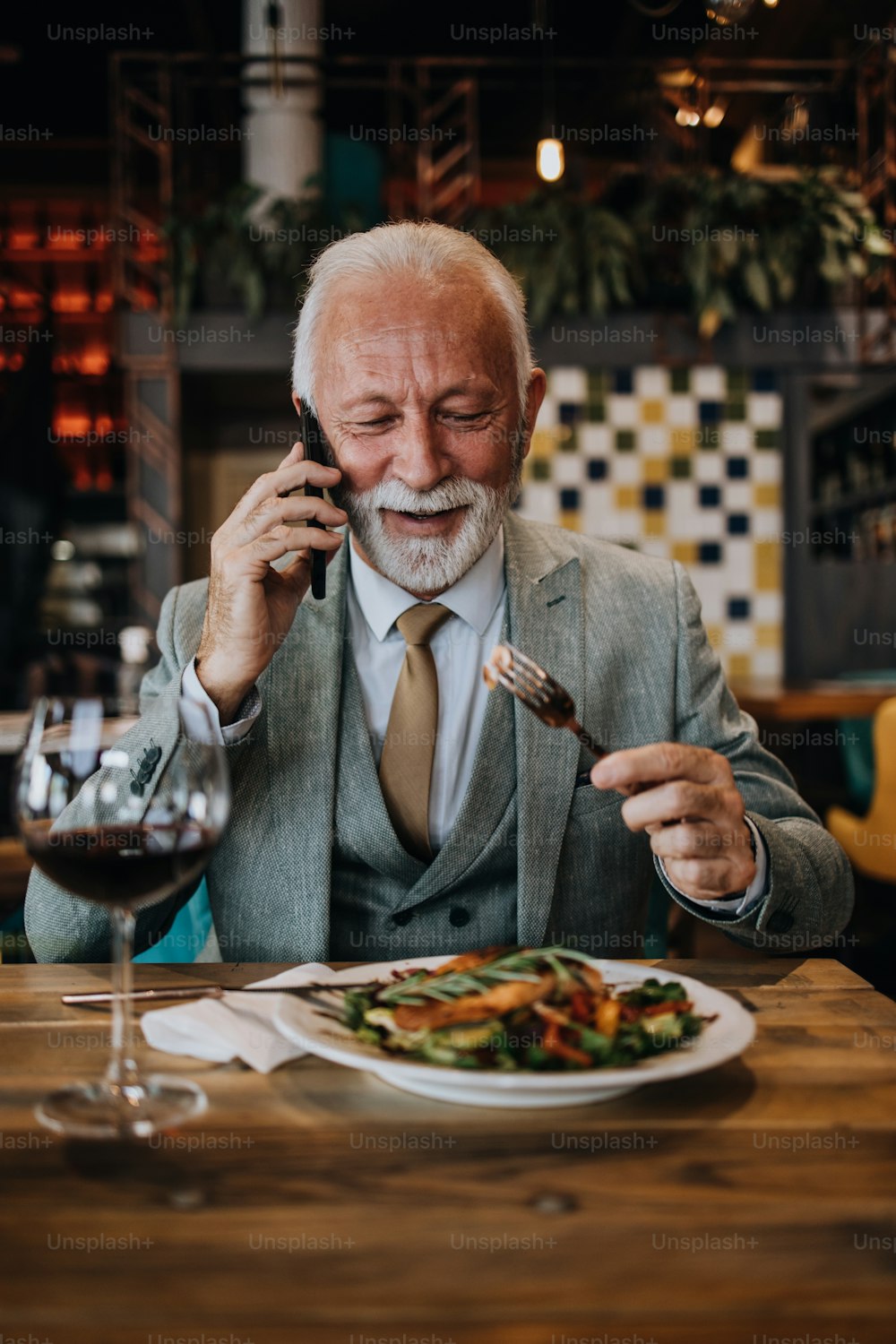 Uomo d'affari anziano felice e bello seduto nel ristorante e in attesa del pranzo. Sta usando lo smartphone e sta parlando con qualcuno. Concetto di stile di vita per anziani d'affari.