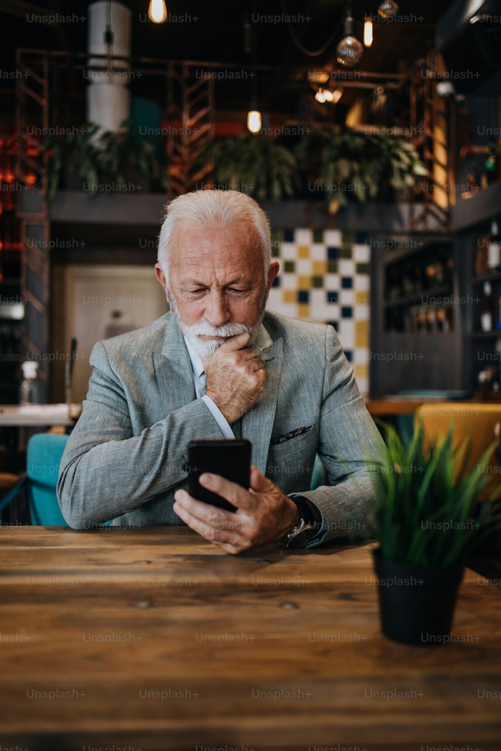 Glücklicher und gutaussehender Geschäftsmann, der im Restaurant sitzt und auf das Mittagessen wartet. Er benutzt sein Smartphone und spricht mit jemandem. Business Senioren Lifestyle-Konzept.