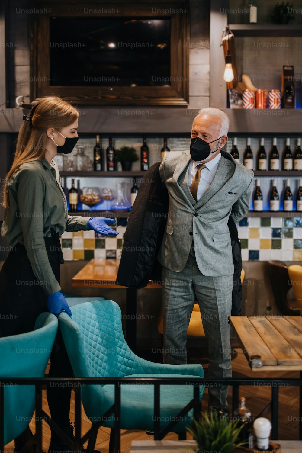 Confident senior businessman standing in exclusive restaurant. He takes off his coat while the waitress helps him. They both wearing protective face masks against virus infection.