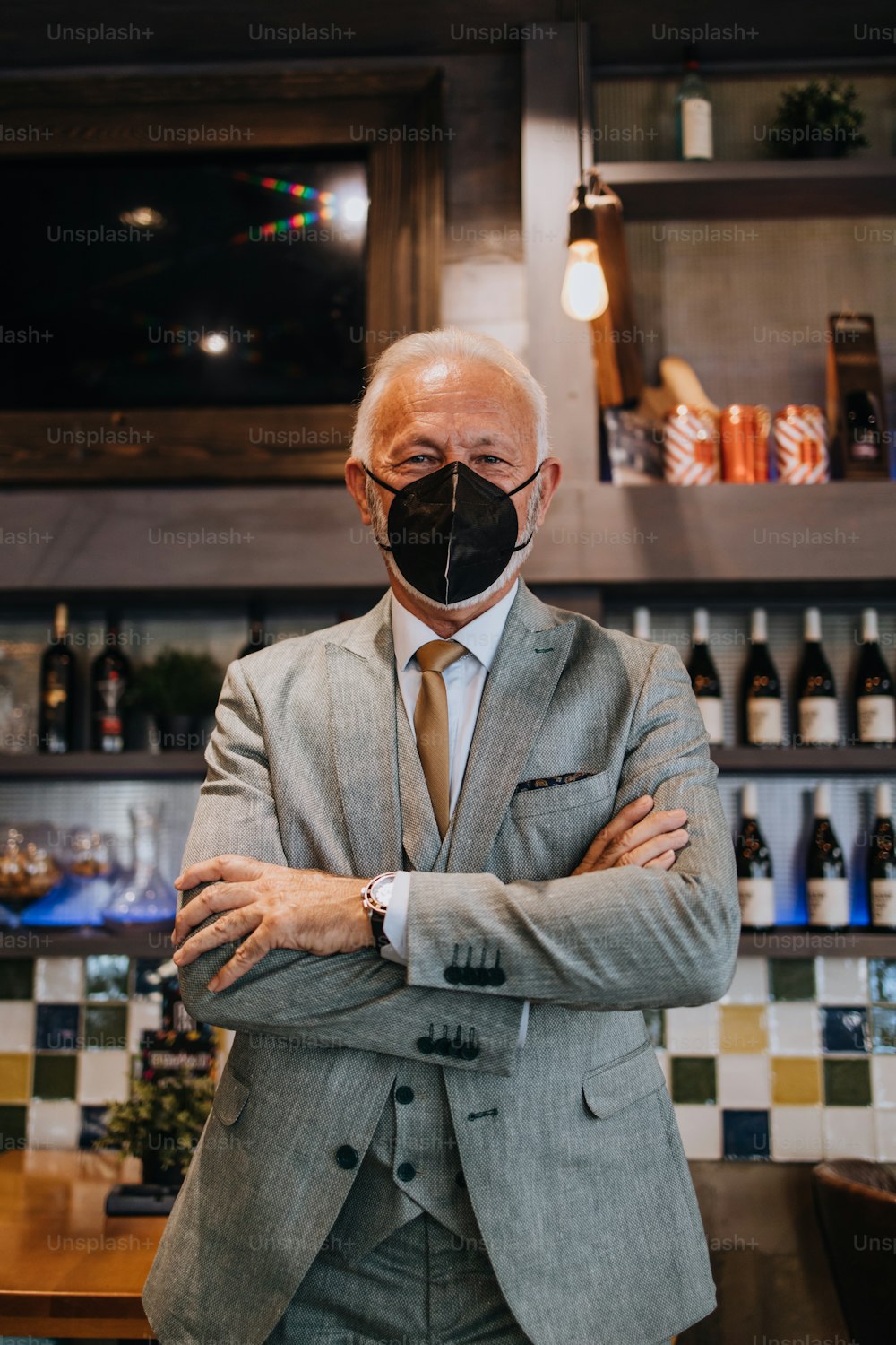 Confident senior businessman standing in exclusive restaurant. He is wearing protective N-95 face mask as protection against virus infection. Coronavirus senior people lifestyle concept.