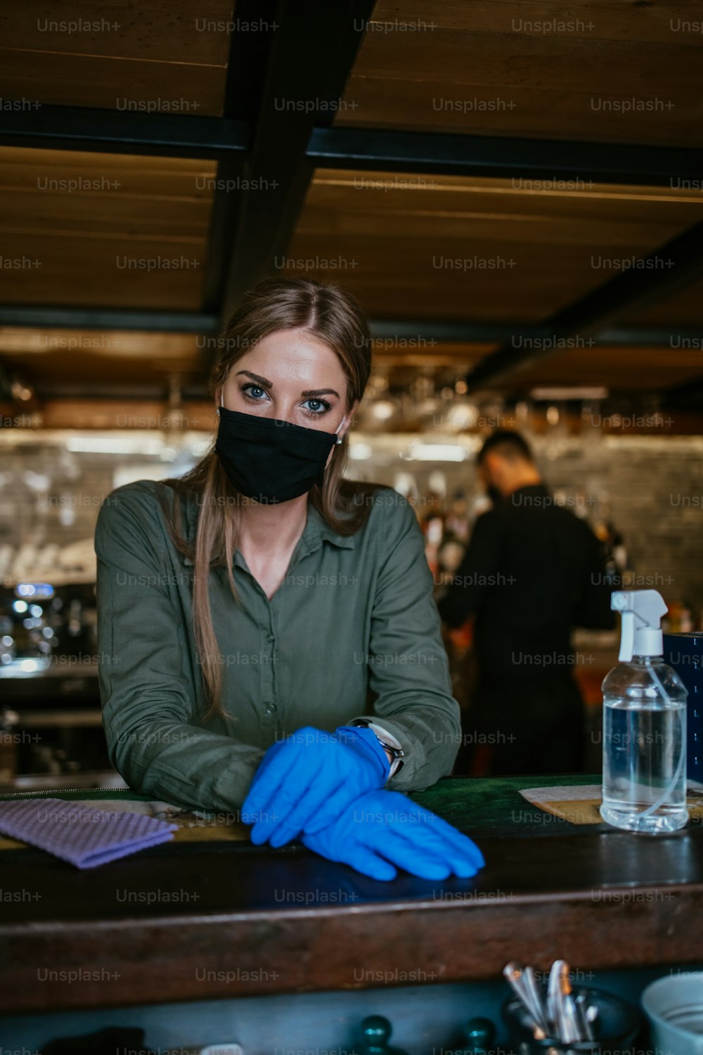 Portrait of a serious waitress standing behind a bar in a nice restaurant. She wears a protective mask and gloves as part of security measures against the Coronavirus pandemic.
