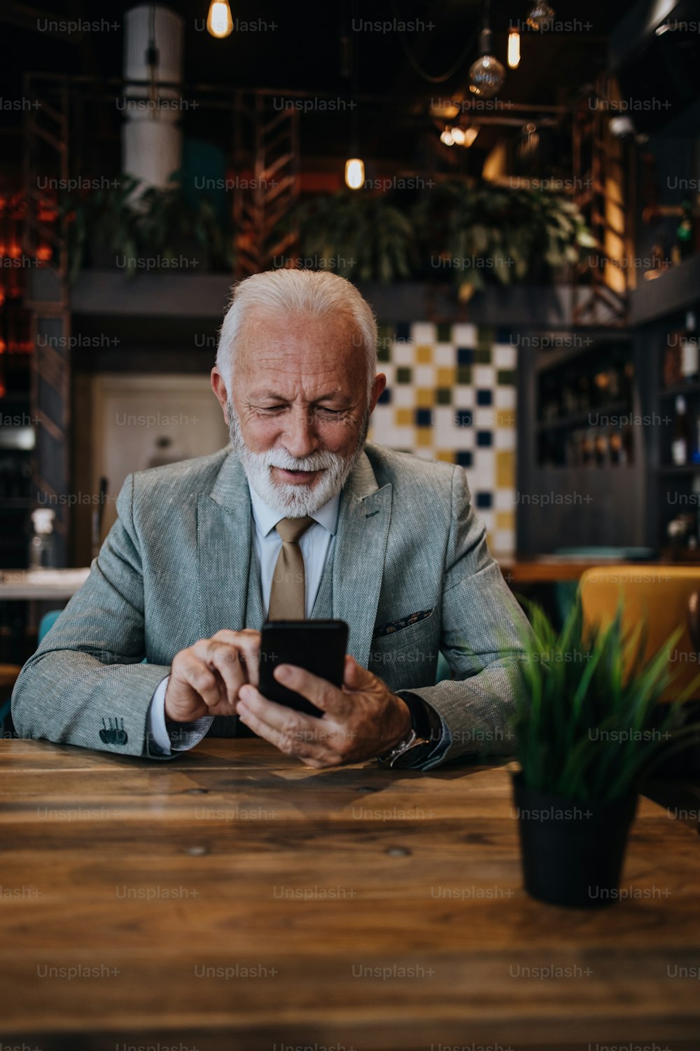 Glücklicher und gutaussehender Geschäftsmann, der im Restaurant sitzt und auf das Mittagessen wartet. Er benutzt sein Smartphone und spricht mit jemandem. Business Senioren Lifestyle-Konzept.