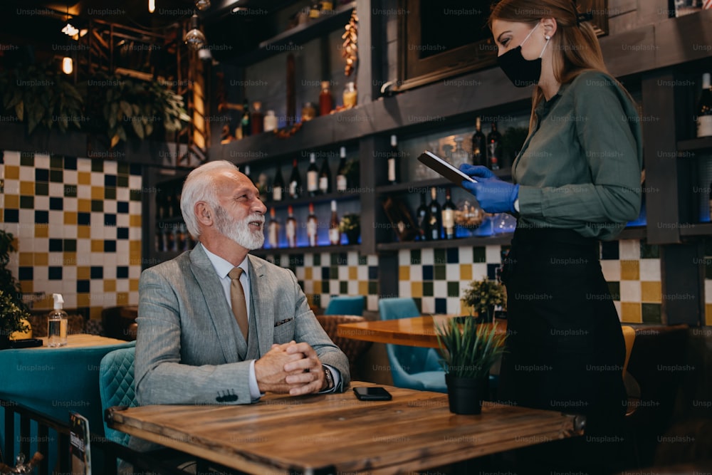 Waitress serves and takes the order from the senior businessman at the restaurant. She wears a protective mask as part of security measures against the Coronavirus pandemic.