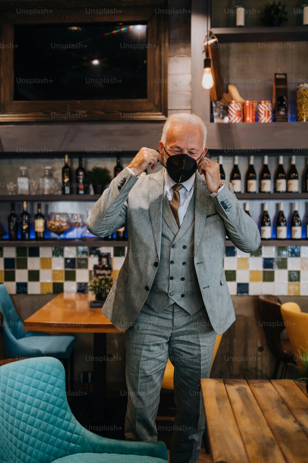 Confident senior businessman standing in exclusive restaurant. He is wearing protective N-95 face mask as protection against virus infection. Coronavirus senior people lifestyle concept.