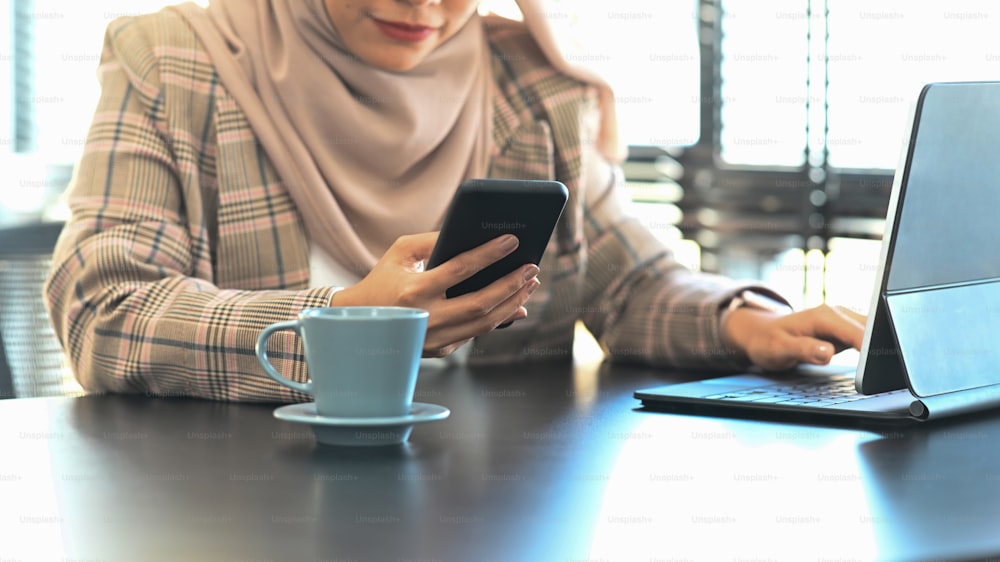 Ausschnittaufnahme einer glücklichen muslimischen Frau, die ein Smartphone benutzt, während sie in ihrem Bürozimmer sitzt.