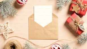 クリスマスの手紙のコンセプト。クラフト紙の封筒、クリスマスプレゼント、木製の装飾、ベージュの背景に松の木の枝に白紙の紙カード。クリスマス招待状のモックアップ。フラットレイ、上面図。