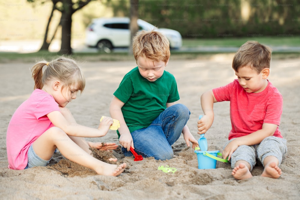 Drei kaukasische Kinder sitzen im Sandkasten und spielen mit Strandspielzeug. Kleine Mädchen und Jungenfreunde haben Spaß zusammen auf dem Spielplatz. Sommer-Outdoor-Aktivität für Kinder. Freizeit Lifestyle Kindheit.
