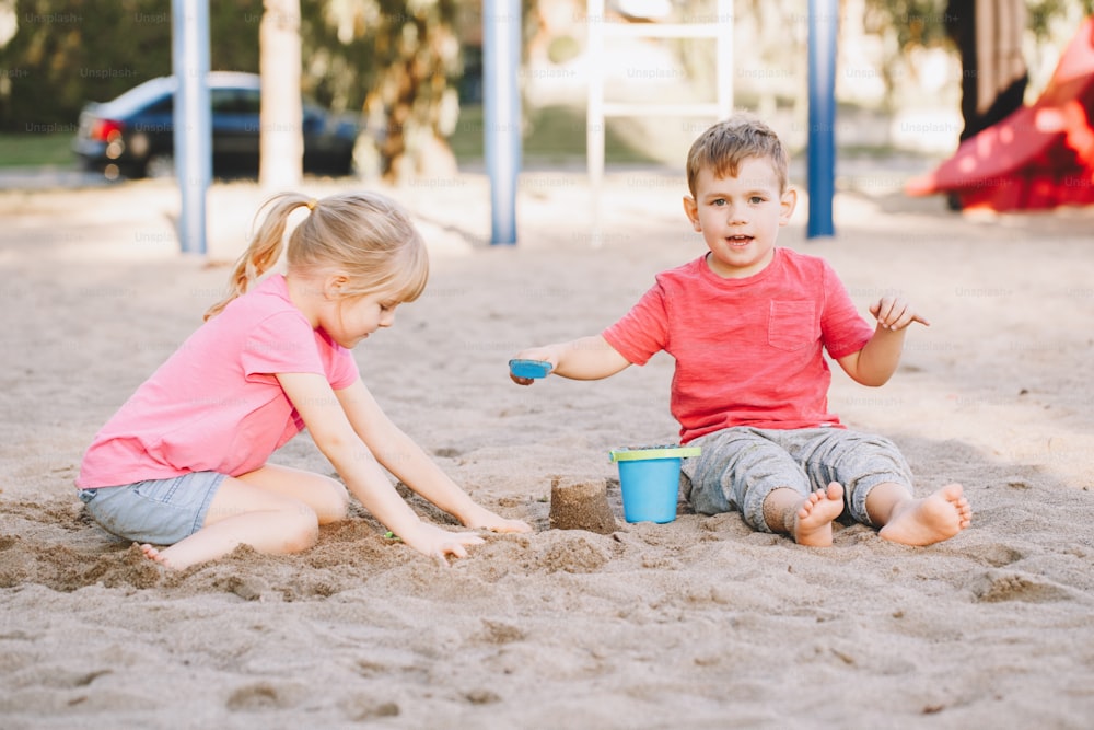 Zwei kaukasische Kinder sitzen im Sandkasten und spielen mit Strandspielzeug. Kleines Mädchen und Freunde haben Spaß zusammen auf dem Spielplatz. Sommer-Outdoor-Aktivität für Kinder. Freizeit Lifestyle Kindheit.