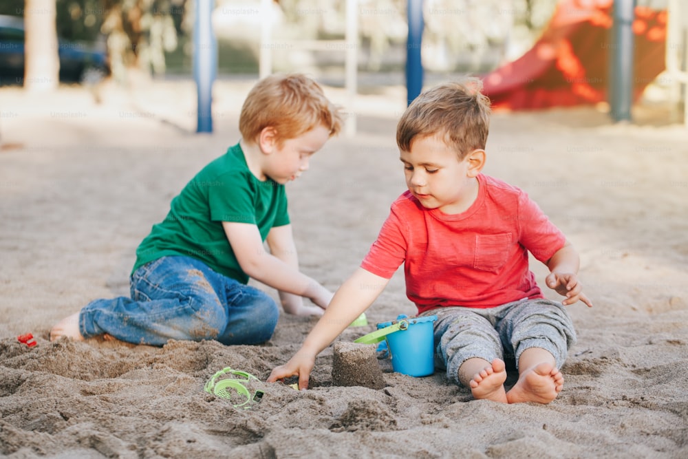 Dos niños caucásicos sentados en un arenero jugando con juguetes de playa. Amigos de niños pequeños divirtiéndose juntos en el patio de recreo. Actividad de verano al aire libre para niños. Tiempo libre, estilo de vida, infancia.