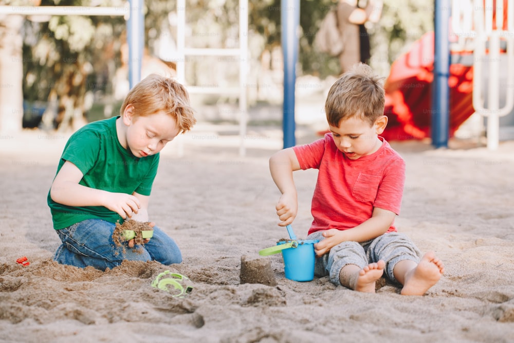 Due bambini caucasici seduti in sabbiera che giocano con giocattoli da spiaggia. Amici dei ragazzini che si divertono insieme al parco giochi. Attività estiva all'aperto per bambini. Tempo libero, stile di vita, infanzia.