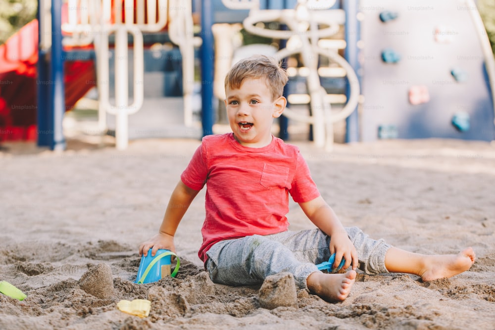 Criança caucasiana sentada em caixa de areia brincando com brinquedos de praia. Baby construindo torta de areia do castelo de areia. Garotinho se divertir no playground. Atividade ao ar livre de verão para crianças. Tempo de lazer estilo de vida infância.
