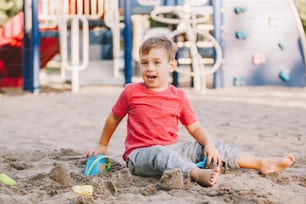 砂場に座ってビーチのおもちゃで遊んでいる白人の子供。ベビービルディングサンドキャッスルサンドパイ。小さな男の子は遊び場で楽しんでいます。子供のための夏の野外活動。余暇、ライフスタイル、子供時代。
