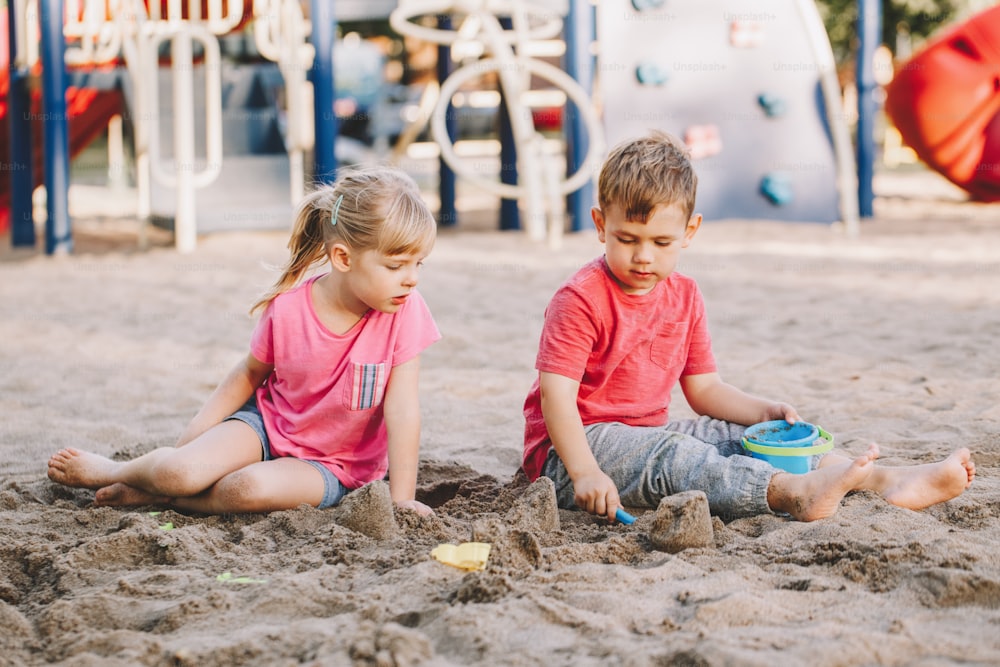 Due bambini caucasici seduti in sabbiera che giocano con giocattoli da spiaggia. Bambina e ragazzi che si divertono insieme nel parco giochi. Attività estiva all'aperto per bambini. Tempo libero, stile di vita, infanzia.