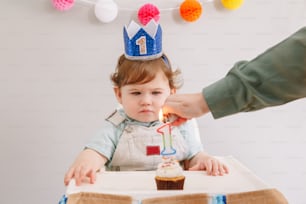 Cute adorable Caucasian baby boy in blue crown celebrating his first birthday at home. Child kid toddler sitting in high chair looking at cupcake dessert. Mother lighting birthday candle on cake.