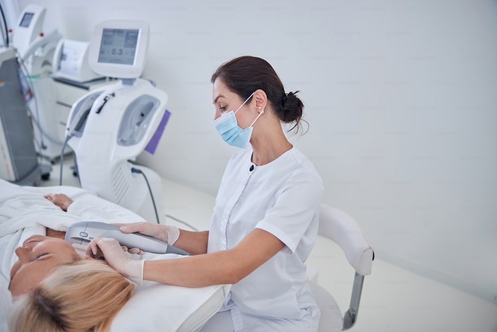 Brunette doctor in medicine mask sitting on a chair while doctor using special cosmetology device for anti age massaging in cosmetological clinic