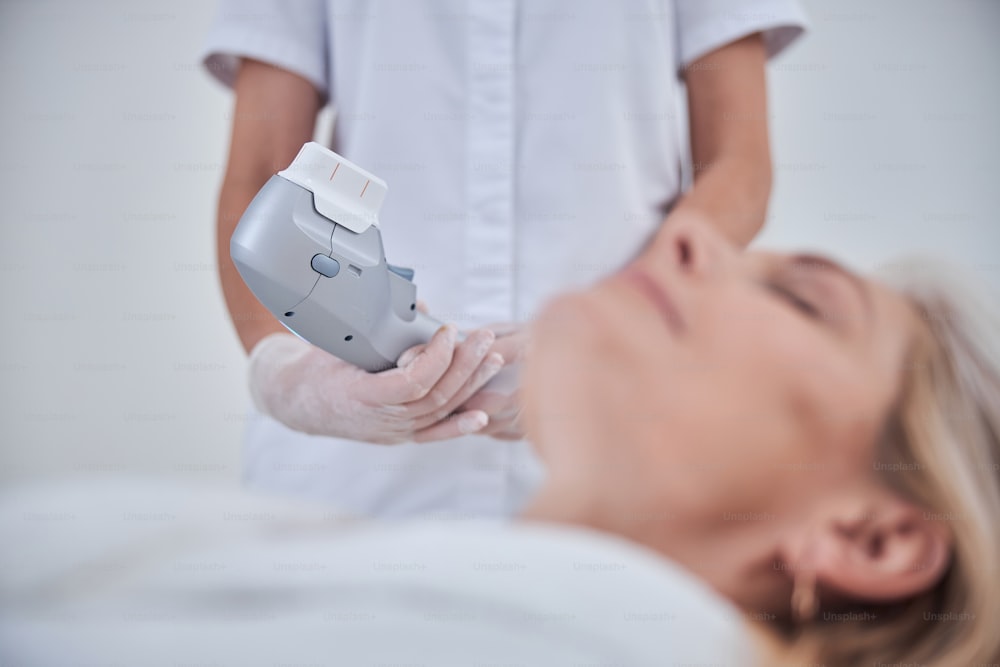 Retrato de perto da máquina de ultrassom moderna nas mãos do médico enquanto o médico em pé perto da paciente do sexo feminino da clínica cosmetológica