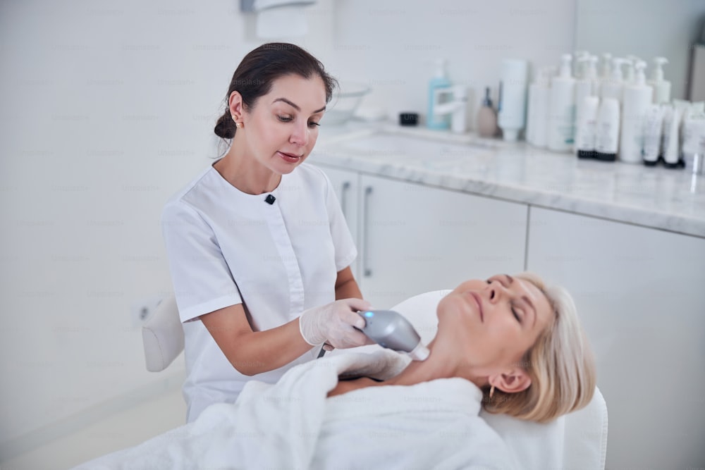 Waist up portrait of elegant stylish blonde Caucasian woman lying on beauty couch while receiving ultrasound anti aging treatment in modern cosmetological