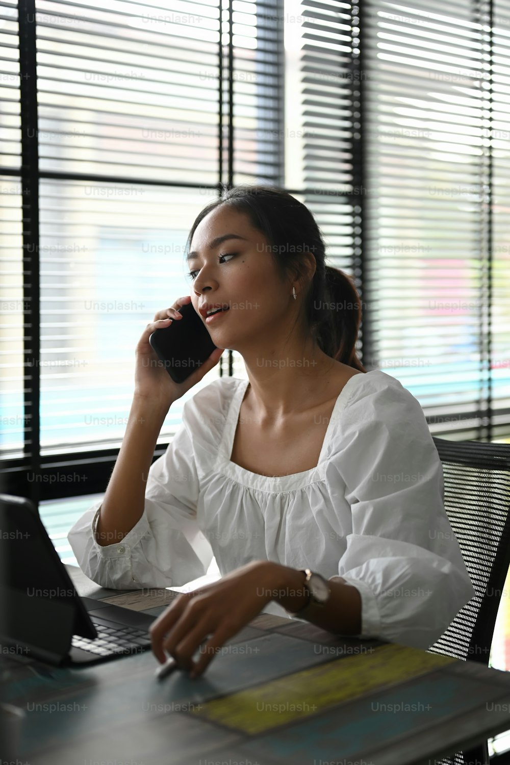 Una joven empleada alegre está hablando por teléfono móvil y usando una tableta de computadora en su escritorio de trabajo.