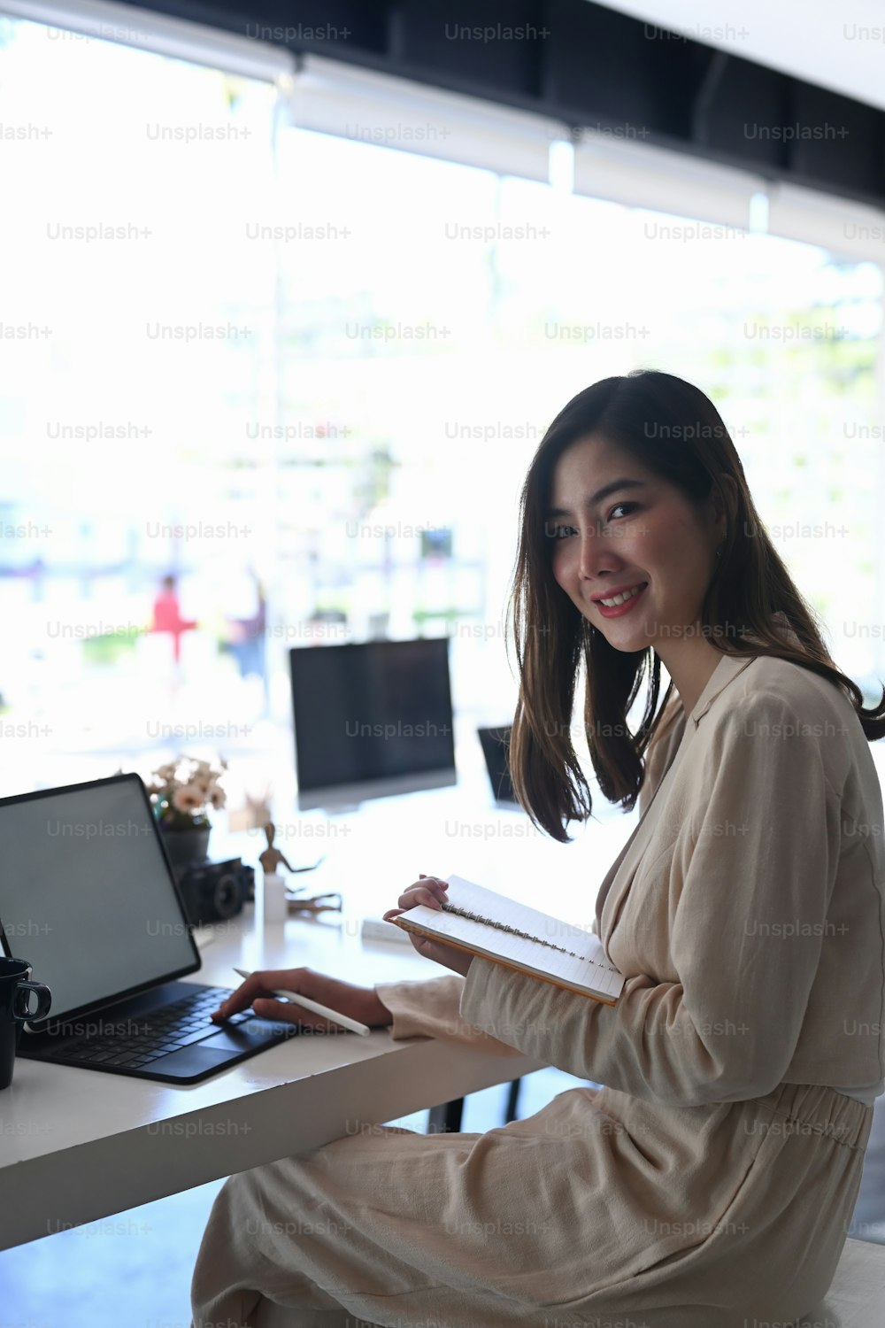 Side view of cheerful businesswoman sitting at her workspace holding notebook and smiling to camera.