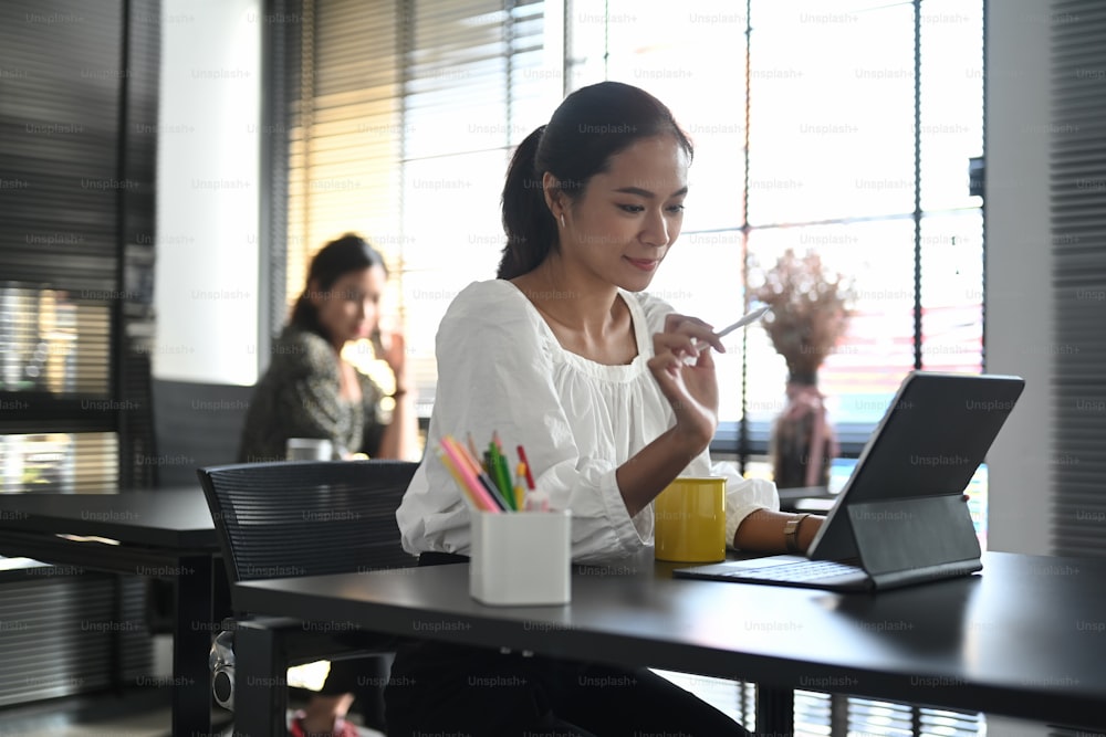 Uma empresária feliz trabalhando em um tablet com seu colega sentado ao fundo.