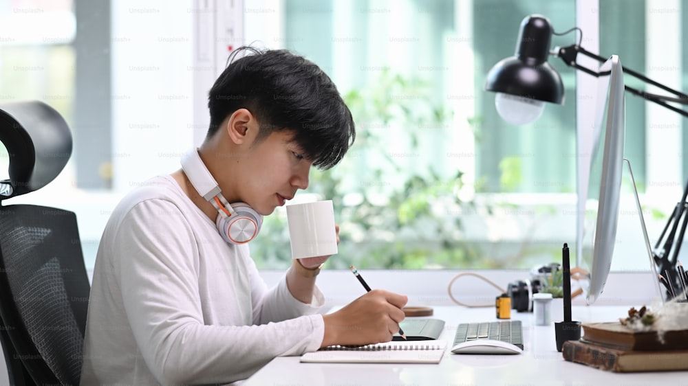 현대적인 사무실에서 커피를 마시는 동안 디지털 태블릿과 스타일러스 펜을 사용하는 헤드폰을 쓴 창의적인 디자이너 남성의 측면 모습.