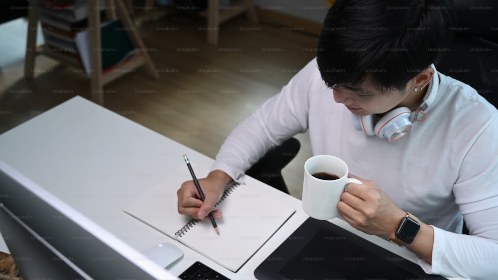 헤드폰을 들고 커피 컵을 들고 창의적인 작업 공간에서 노트북에 글을 쓰는 캐주얼한 남자.