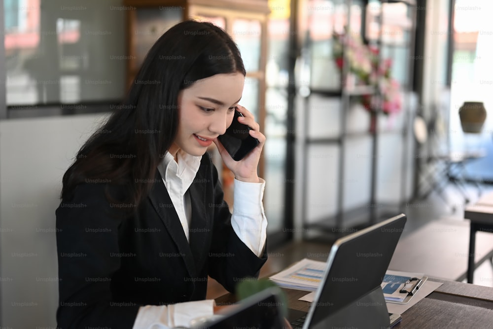 Une belle jeune femme en costume noir parlant sur un téléphone portable et utilisant une tablette alors qu'elle était assise dans la salle de bureau.