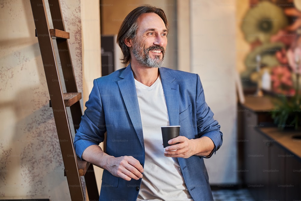 Foto dalla vita in su di un allegro uomo bruno che indossa un blazer mentre tiene in mano una tazza di caffè e sorride