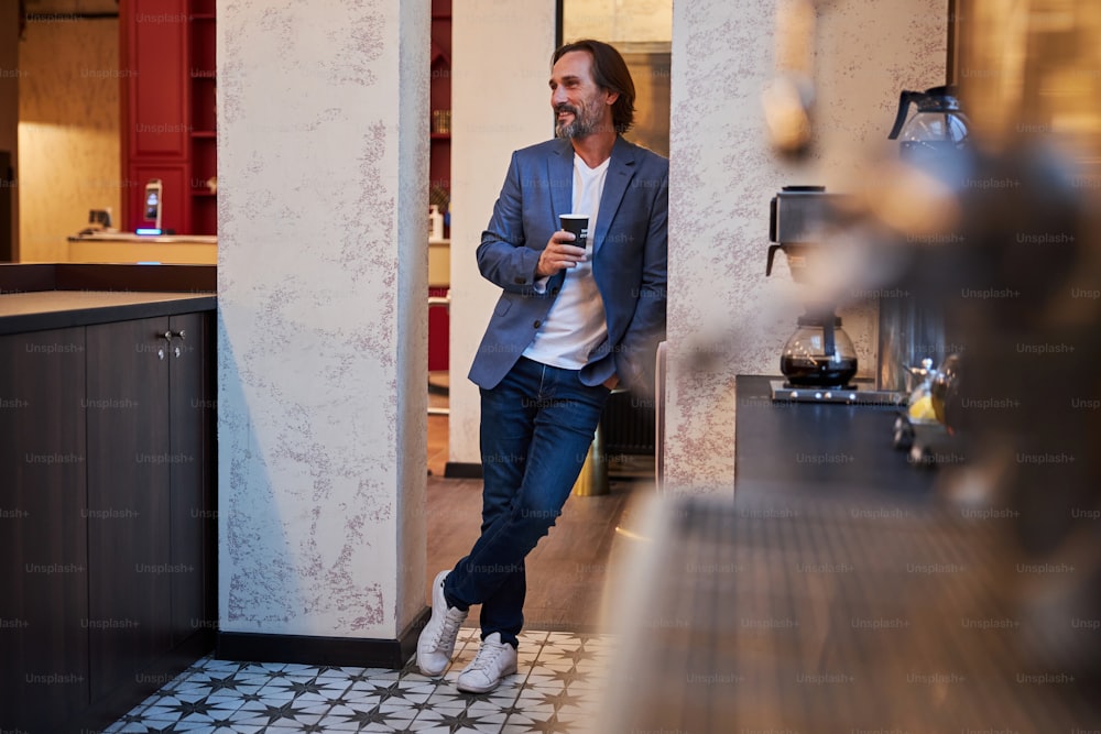 Gentiluomo sorridente rilassato in abiti eleganti-casual appoggiato al muro mentre tiene in mano una tazza di caffè