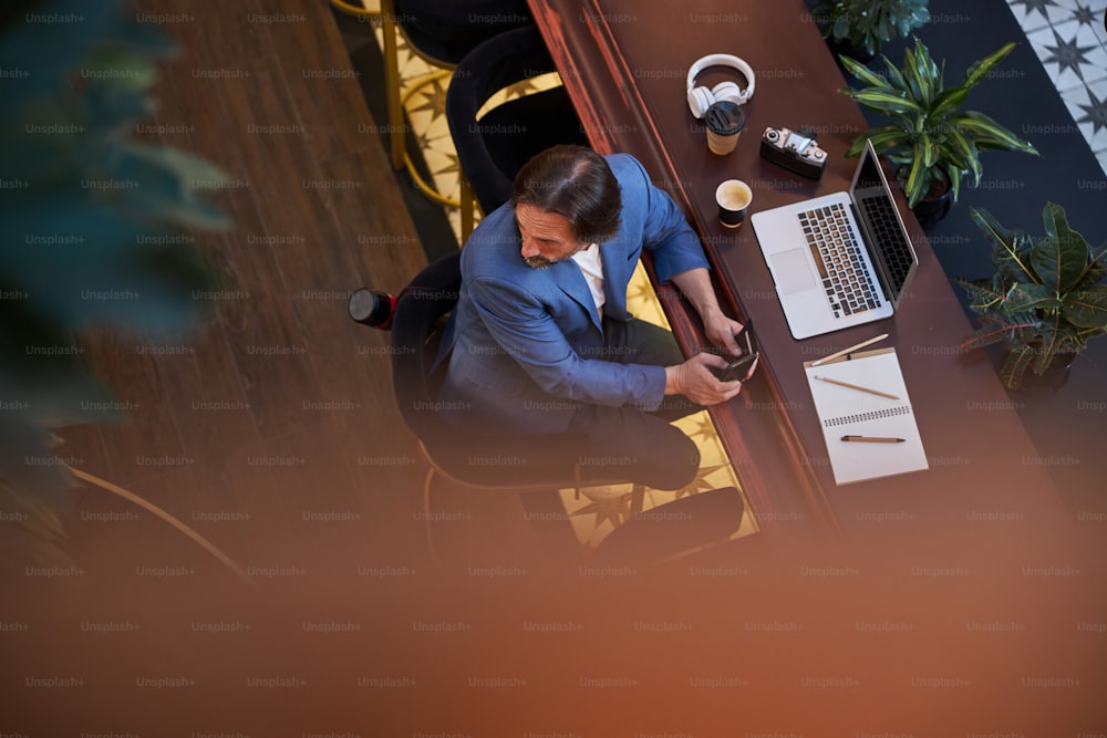 Foto dall'alto di un uomo d'affari in viaggio che gira la testa mentre ha la sua attrezzatura da lavoro disposta su un lungo tavolo marrone