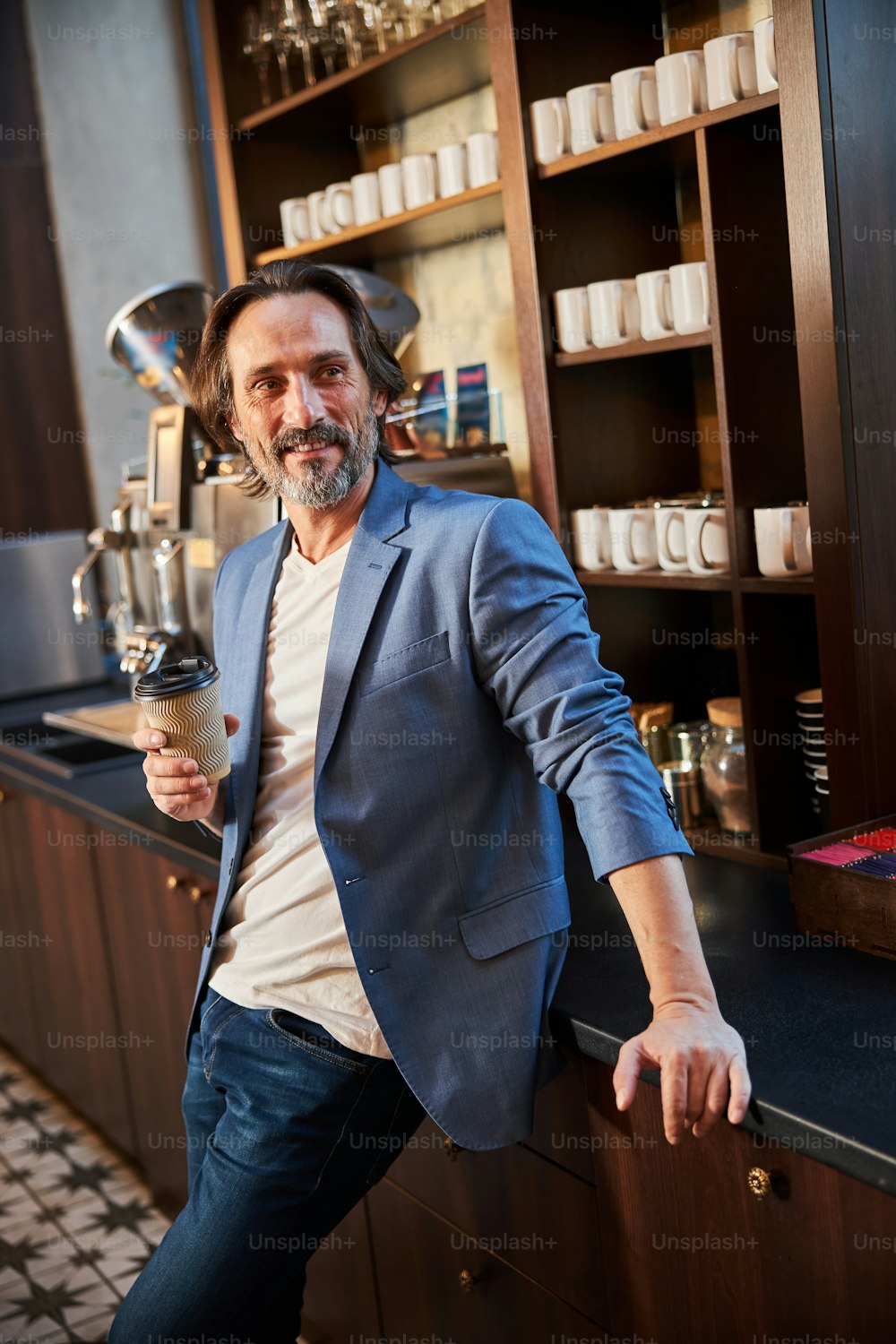 Approachable middle-aged man with paper cup in hand leaning on a coffee bar and looking to the side
