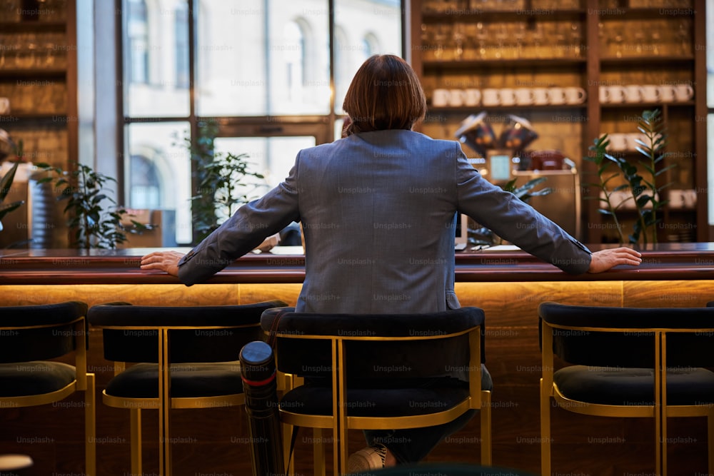 Rear view photo of a brunette man in smart suit sitting and chilling at bar counter of a fancy hotel
