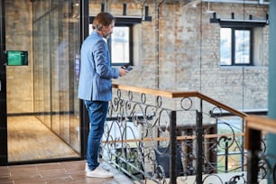 Full-length photo of tall handsome man standing near staircase and looking at his smartphone