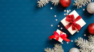 빨간 리본 활, 공 장식 및 색종이가 있는 진한 파란색 배경에 소나무 가지가 있는 흰색 선물 상자. 크리스마스 선물 개념입니다. 플랫 레이, 평면도.